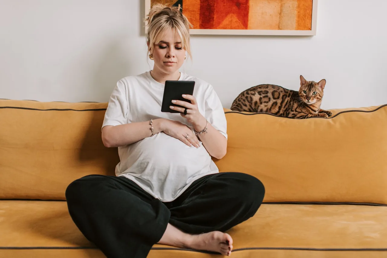 A pregnant woman sitting cross-legged on a sofa with a Bengal cat resting on the backrest behind her.