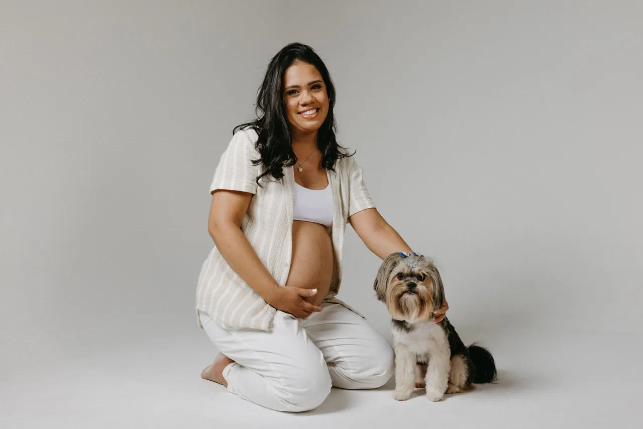 A smiling pregnant woman kneeling on the floor beside her small dog.