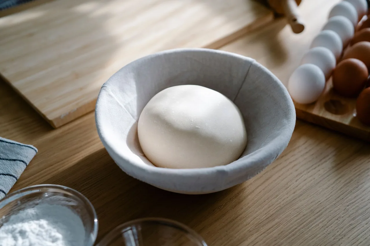 A ball of raw yeast dough resting in a cloth-lined bowl on a kitchen counter, with eggs and flour nearby.