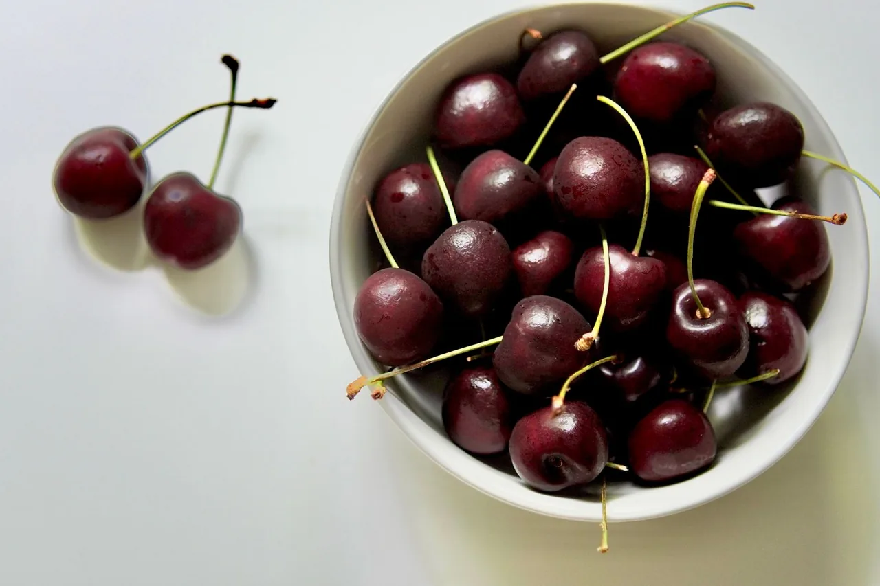 Un tazón blanco de cerezas rojo oscuro con tallos, y dos cerezas sueltas al lado.
