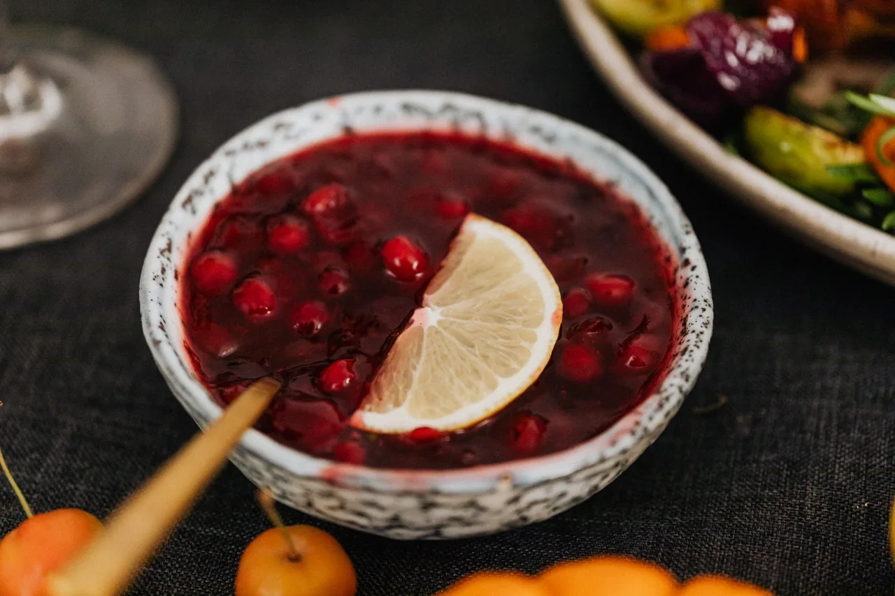 A speckled ceramic bowl of whole-berry cranberry sauce with a lemon slice and a gold spoon, on a dark tablecloth beside a plate of roasted vegetables.