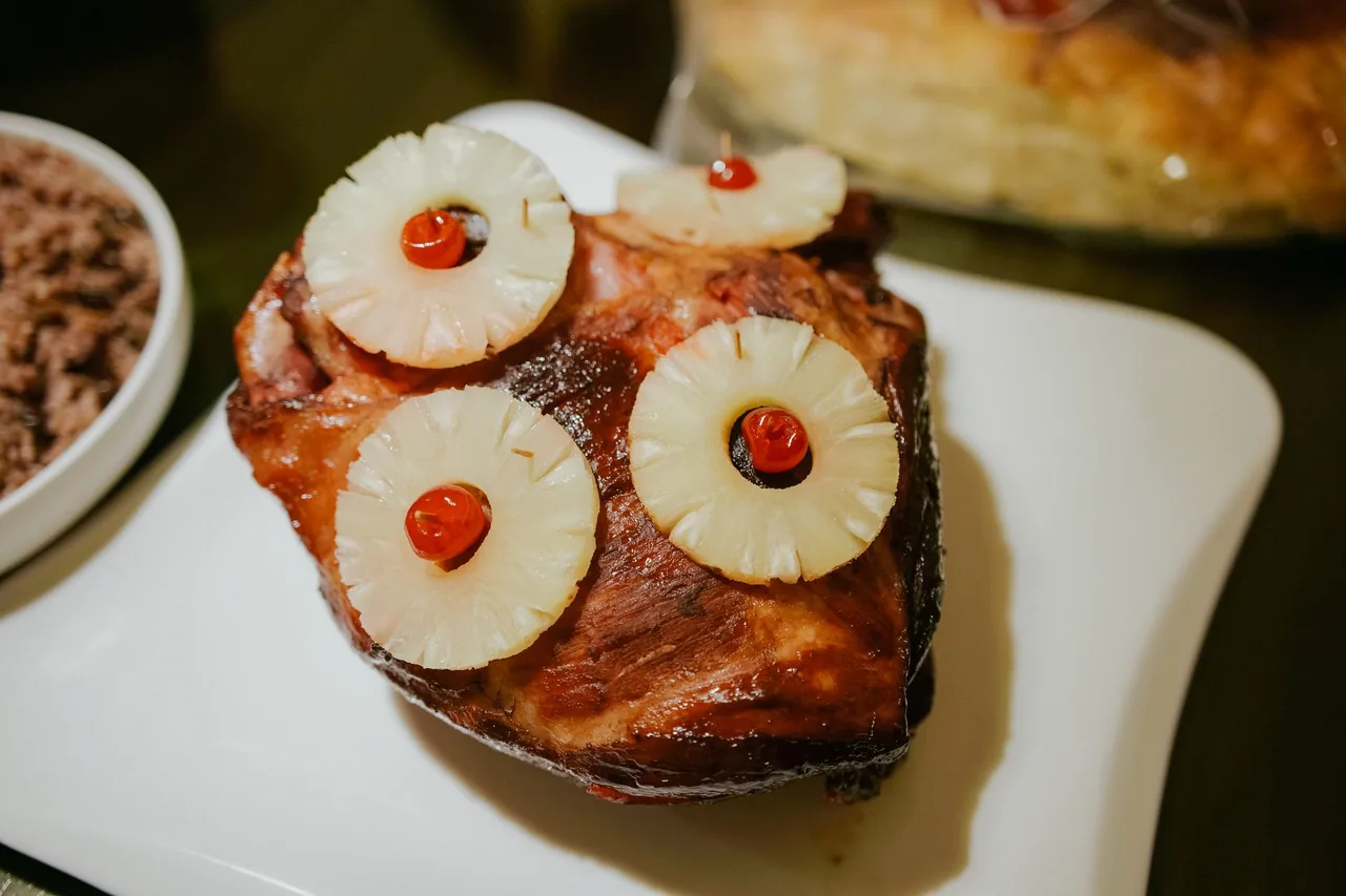 A glazed baked ham on a white platter, decorated with pineapple rings and red cherries, with a side dish blurred in the background.