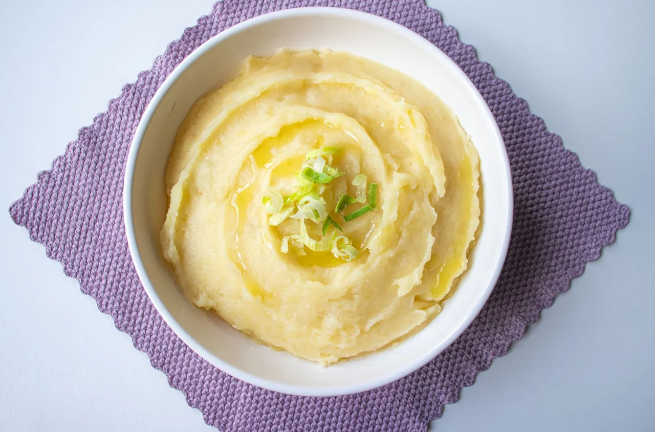 A white bowl of creamy mashed potatoes swirled on a purple woven mat, topped with sliced green onion and a drizzle of melted butter or oil.