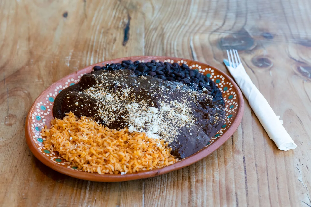 Un plato de mole oscuro con ajonjolí encima, con arroz y frijoles negros.