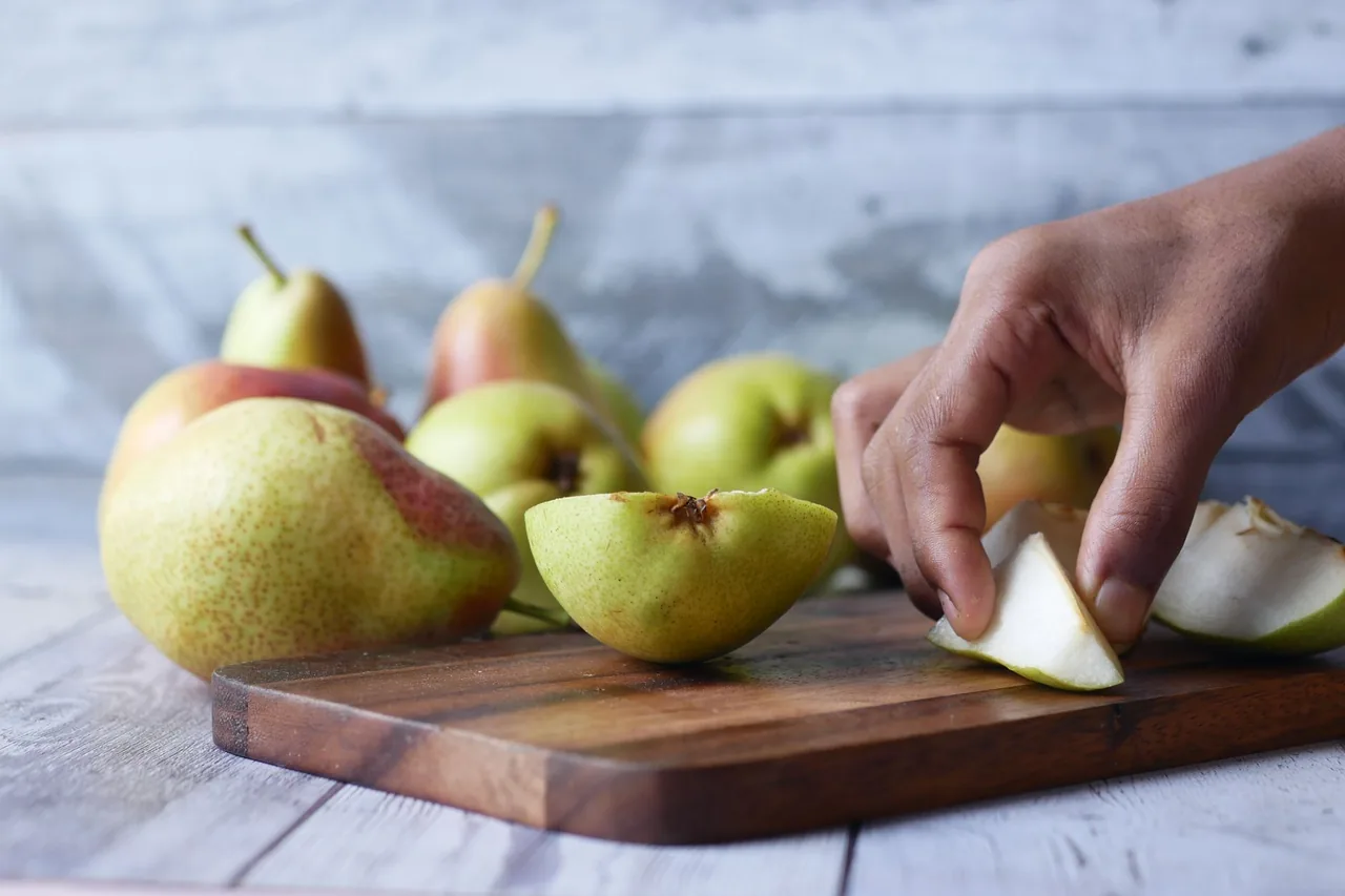 A hand slicing a pear on a wooden board, with whole pears behind it.