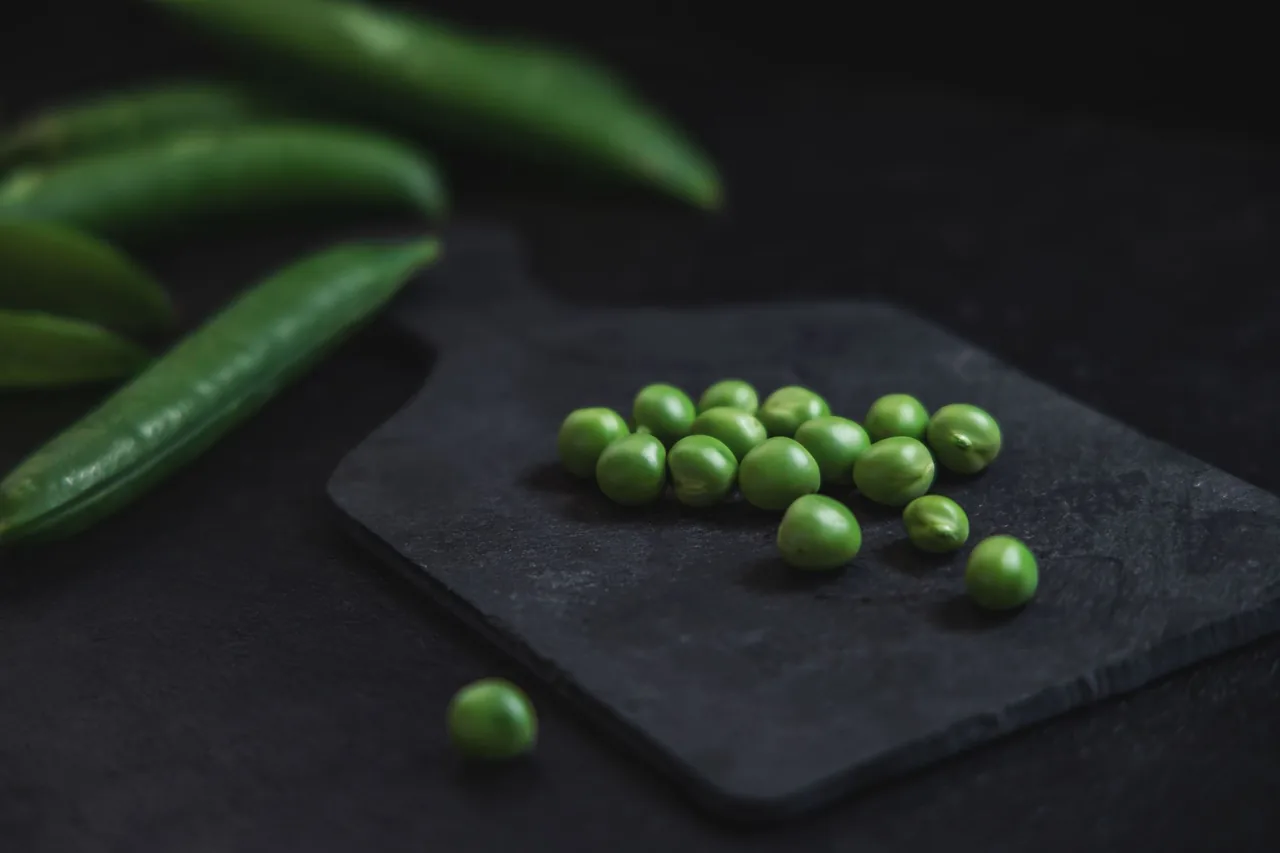Chícharos verdes pelados sobre una tabla oscura, con vainas al lado.