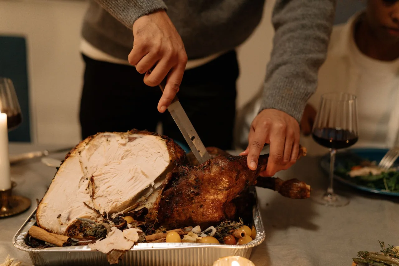 A person carving a whole roast turkey in a foil pan on a set holiday table, with thick slices of white breast meat falling away from the browned skin.