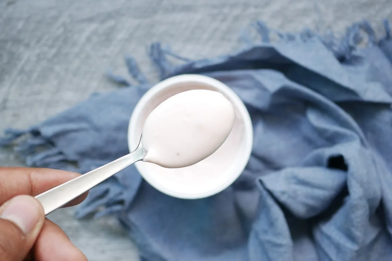 A spoon lifting plain white yogurt from a small white bowl on a blue cloth.