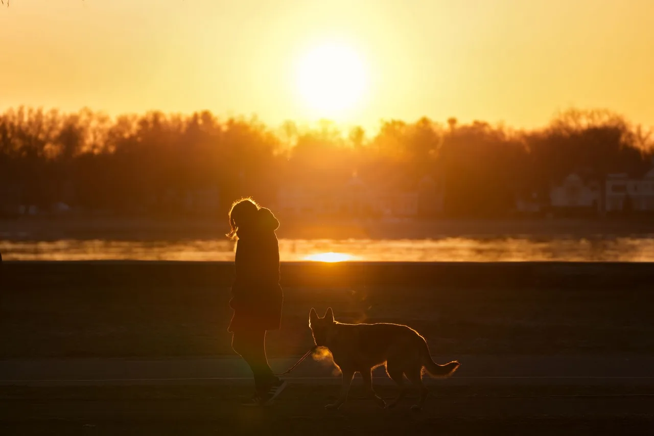 A woman in a winter coat walking a German Shepherd on a leash along a riverside path at sunset.