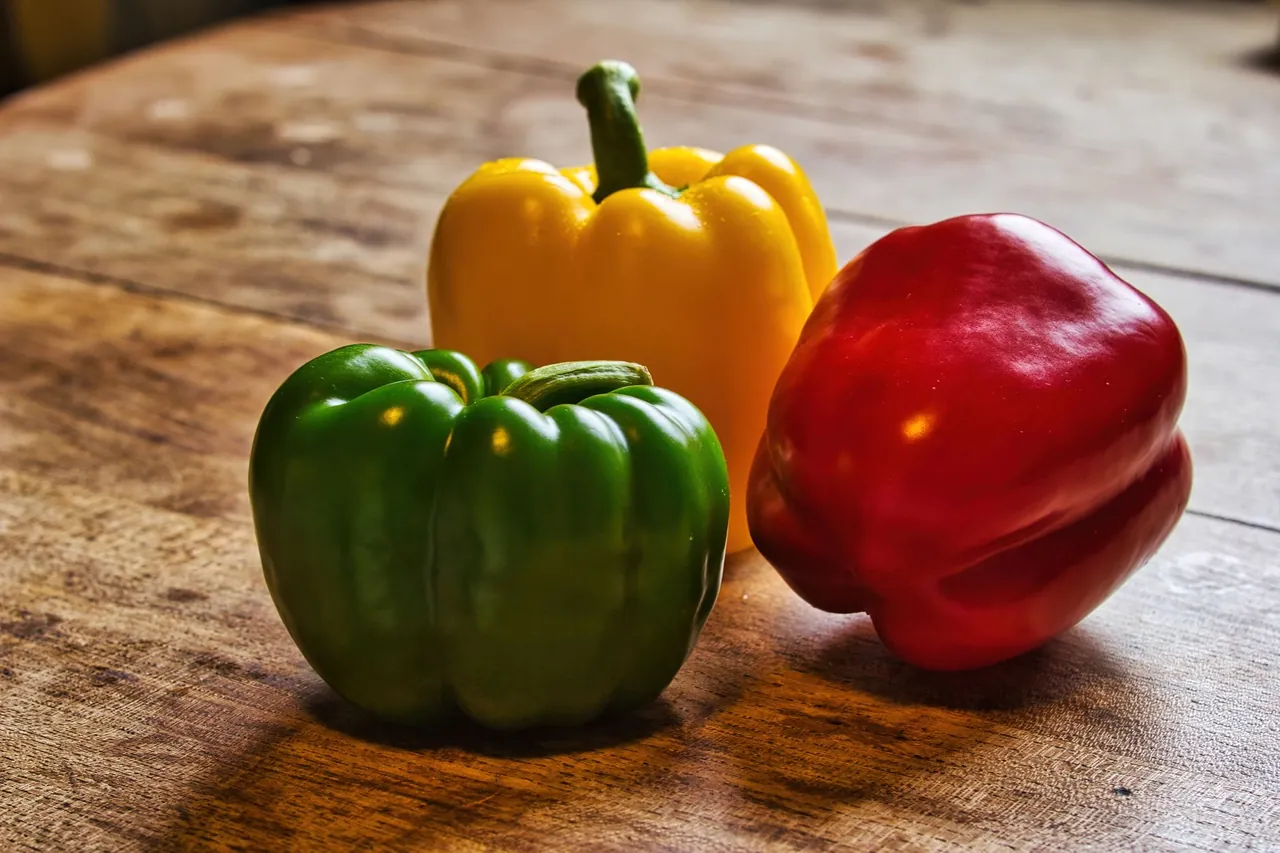 A green, a yellow and a red bell pepper on a wooden table.