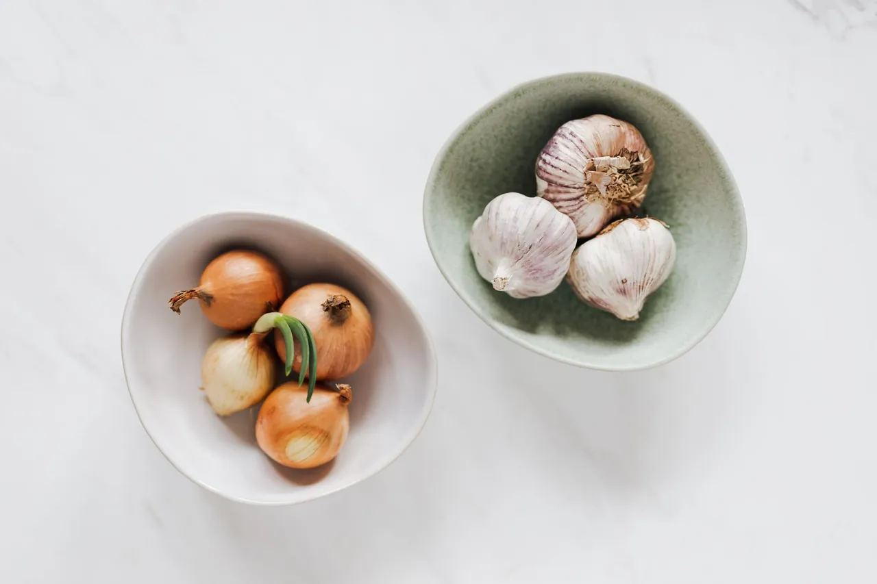 Two white bowls, one with onions and one with garlic bulbs.