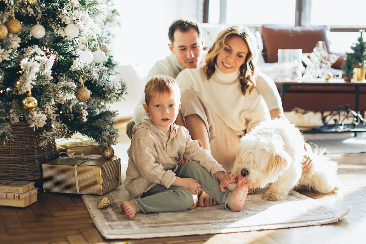 Una familia sentada en el piso junto a un árbol de Navidad, con un perro blanco y peludo al lado de un niño pequeño.