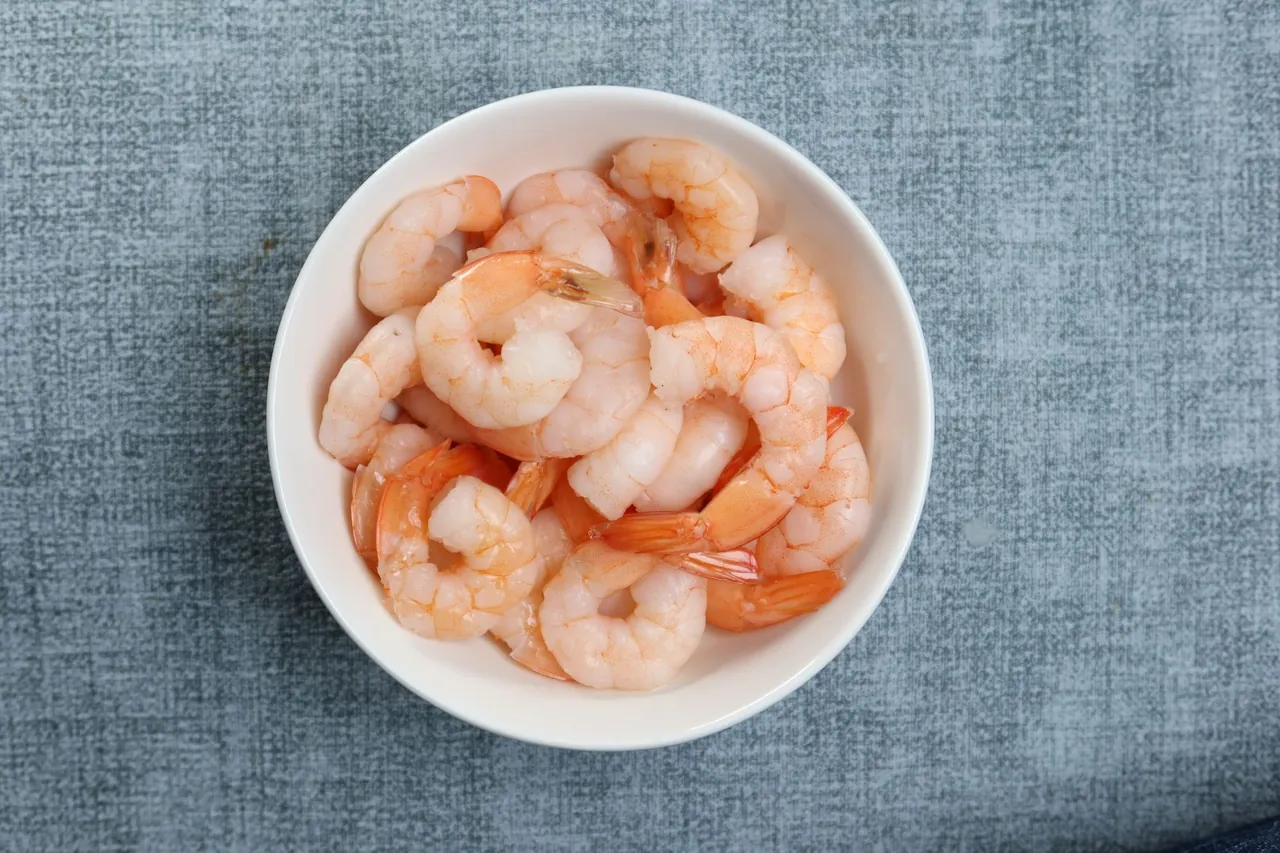 A white bowl of cooked, peeled shrimp, some with tails on, on a blue cloth.