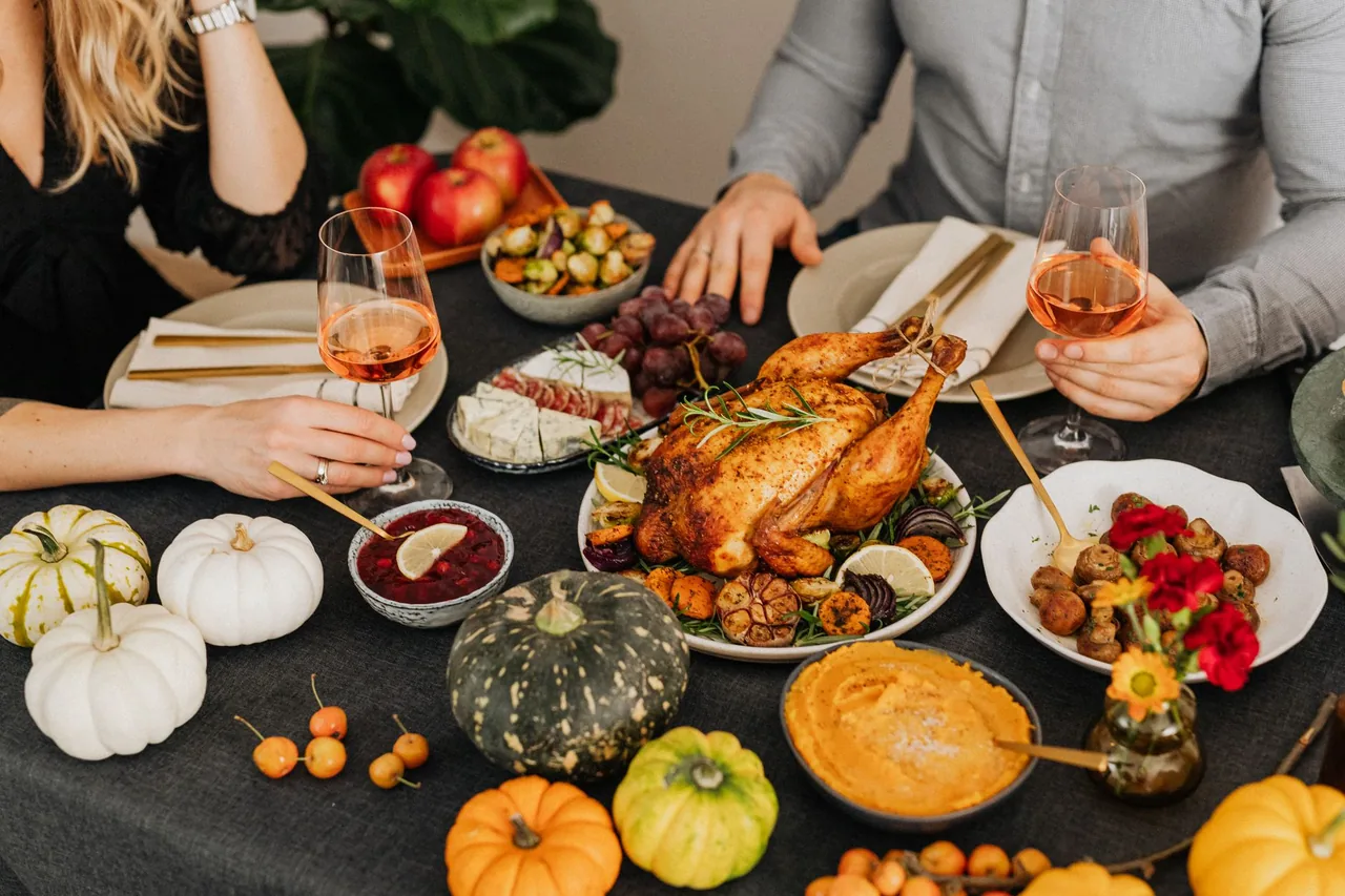 A holiday dinner table set with a roast turkey on a platter beside roasted garlic, a bowl of red grapes, a cheese plate, cranberry sauce, mashed squash, and small decorative pumpkins.